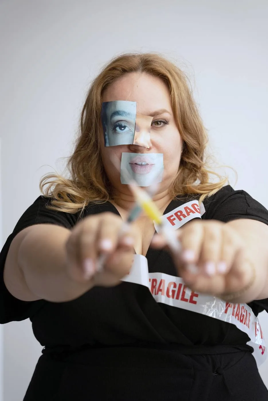 A woman holding out her hands, with cut-out photographs of other people's eyes and lips taped over her face and tape marked FRAGILE across her shoulder